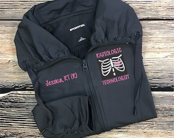 May include: A dark gray zippered jacket with a pink embroidered design of a ribcage with a heart inside. The text "Radiologic Technologist" is embroidered below the design. The text "Jessica, RT (R)" is embroidered on the left sleeve.