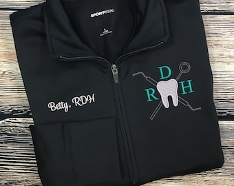 Op de afbeelding: Zwarte ritsjas met een groen en grijs geborduurde tand, tandheelkundige instrumenten en de letters "DRH". De tekst "Betty, RDH" is geborduurd op de linkerzijde van de jas.