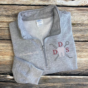 May include: Gray zippered sweatshirt with a white embroidered design of a tooth, dental tools, and the letters "DDS". The text "Dr. Kara Miller, DDS" is also embroidered on the sweatshirt.