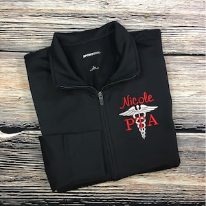 May include: Black zippered jacket with a red embroidered name "Nicole" and a silver medical symbol with the letters "PTA" below it.