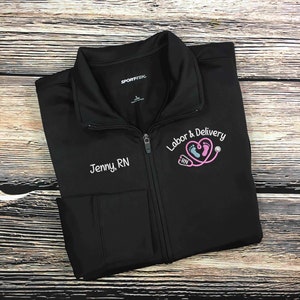 May include: Black zippered jacket with a pink embroidered heart and the text "Labor & Delivery RN" on the right side. The text "Jenny, RN" is embroidered on the left side of the jacket.