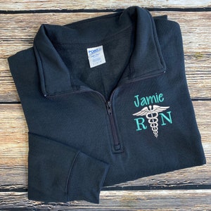 May include: Black 1/4 zip sweatshirt with a teal embroidered medical symbol and the text "Jamie RN".
