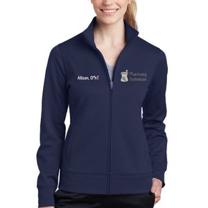 May include: Navy blue zip-up jacket with the text "Allison, CPhT" and a pharmacy technician logo. The jacket has a high collar and two front pockets. The model is wearing black pants.