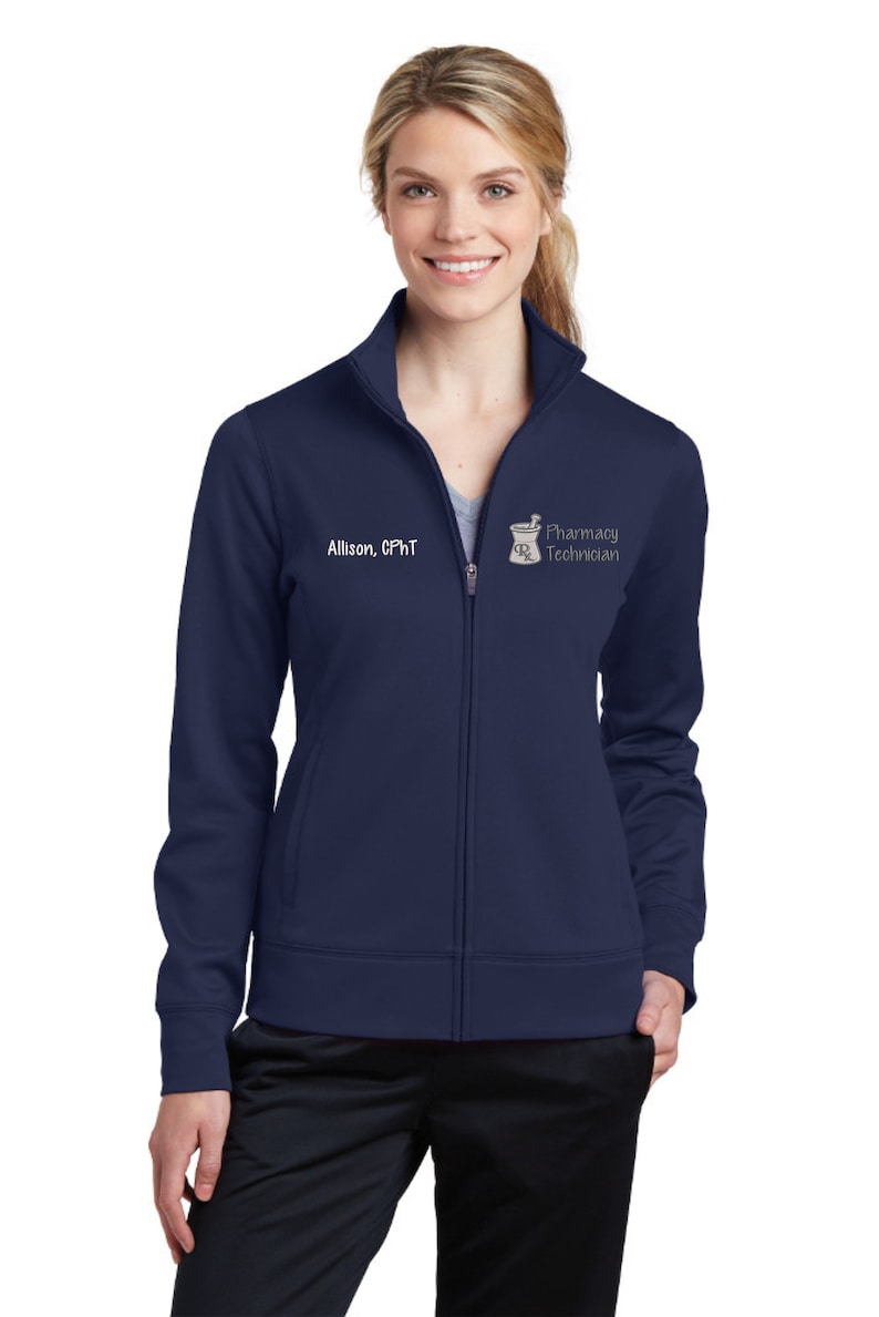 May include: Navy blue zip-up jacket with the text "Allison, CPhT" and a pharmacy technician logo. The jacket has a high collar and two front pockets. The model is wearing black pants.