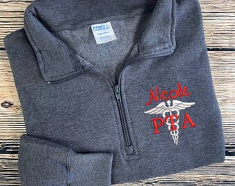 May include: A dark gray 1/4 zip sweatshirt with a white embroidered medical caduceus symbol and the text "Nicole PTA" in red.