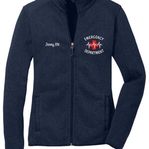 May include: A navy blue fleece zip-up jacket with a white embroidered logo that says "Emergency Department" with a red cross and a heartbeat graphic. The jacket also has a white embroidered name tag that says "Jenny, RN".