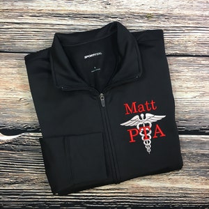 May include: Black zippered jacket with a red embroidered name "Matt" and a silver medical symbol with the letters "PTA" underneath.