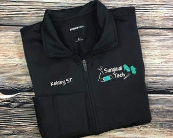 May include: Black zippered jacket with a pocket on the left side. The jacket has the text "Kelsey, ST" embroidered on the pocket and "SPORT-TEK" on the upper left side. The right side of the jacket has a teal green embroidered design of surgical tools, a bandage, and the text "Surgical Tech".