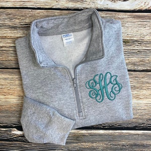 May include: A gray quarter-zip sweatshirt with a teal monogram embroidered on the front. The sweatshirt is made by Port Company.