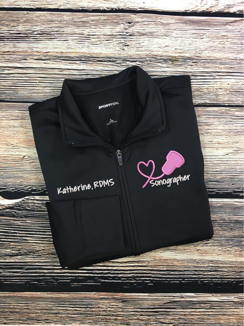 May include: Black zippered jacket with a pink heart and stethoscope graphic and the text "Sonographer" and "Katherine, RDMS".