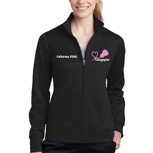 K&ouml;nnte beinhalten: Schwarze Rei&szlig;verschlussjacke mit einem wei&szlig;en gestickten Herz- und Stethoskop-Design. Der Text "Sonographin" ist unter dem Design gestickt. Der Text "Katherine, RDMS" ist auf der linken Seite der Jacke gestickt.