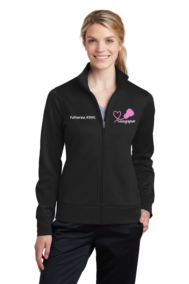 K&ouml;nnte beinhalten: Schwarze Rei&szlig;verschlussjacke mit einem wei&szlig;en gestickten Herz- und Stethoskop-Design. Der Text "Sonographin" ist unter dem Design gestickt. Der Text "Katherine, RDMS" ist auf der linken Seite der Jacke gestickt.