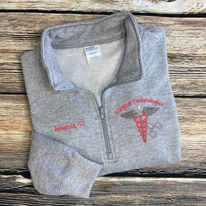 May include: Gray zippered sweatshirt with the text "Surgical Technologist" and a red medical symbol embroidered on the front. The text "Amanda ST" is embroidered on the left sleeve.