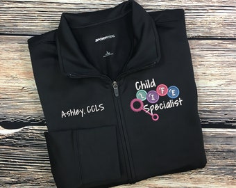 Może przedstawiać: Czarna kurtka zapinana na zamek błyskawiczny z napisem "Child Life Specialist" i kolorowym wzorem koła zębatego wyszytym z przodu. Napis "Ashley, CCLS" jest wyszyty po lewej stronie kurtki.