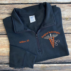 May include: Black quarter-zip sweatshirt with the text "William, ST" embroidered in orange. The sweatshirt also features the words "Surgical Technologist" and a medical symbol with wings and scissors.