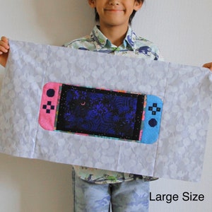 May include: A white fabric pillow with a large, colorful embroidered image of a video game console. The console is pink and blue with a black screen showing a dark blue and purple forest scene. The text "Large Size" is printed in black at the bottom of the pillow.