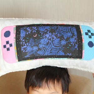 May include: A white pillow with a blue, pink, and black design of a video game controller. The controller has a screen with a blue and black floral pattern.