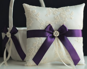 May include: White satin ring pillow with a purple ribbon and a pearl-like button. The pillow is decorated with lace and is sitting on a white basket.