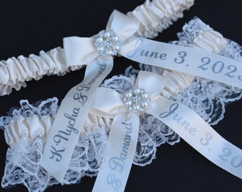 May include: Two ivory elastic garters featuring white lace trim and a central satin bow adorned with a circular pearl and rhinestone embellishment. Each garter includes a satin ribbon printed with the text K. Nycha & Damond and June 3, 2023.
