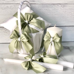 May include: A wedding ceremony set with white satin ring pillows, a white satin ring bearer basket with a sage green bow, and two white unity candles with sage green bows.