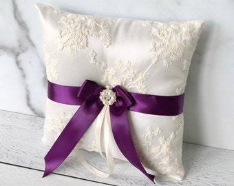May include: A white satin ring pillow with a delicate floral embroidery design. It features a purple satin ribbon tied into a bow with a pearl and rhinestone embellishment.