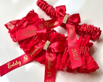 May include: Two red garters with gold lettering. One garter says "Eddy & Nena" and the other says "October 14, 2023".