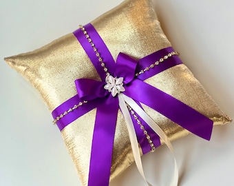 May include: A gold pillow with a purple satin ribbon and a white flower brooch. The pillow is decorated with a string of rhinestones.