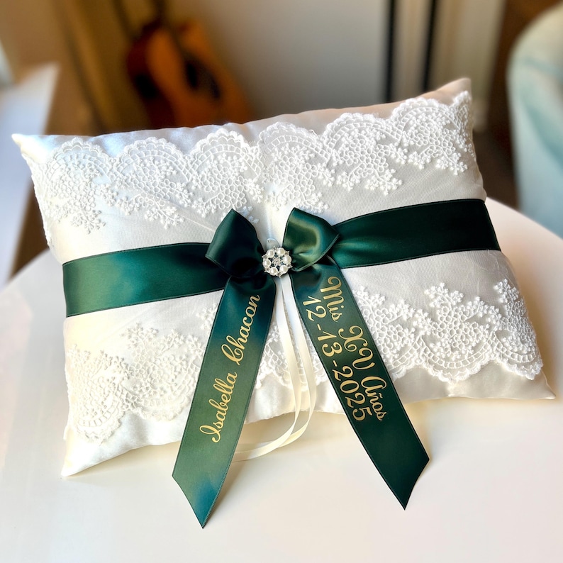Peut inclure: Un coussin d'alliances en satin blanc avec une bordure en dentelle et un ruban vert fonc&eacute;. Le ruban est orn&eacute; d'un bijou en strass et de deux rubans verts avec le texte dor&eacute; "Isabella Chacon" et "Mis XV A&ntilde;os 12-13-2025".
