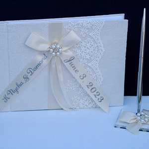 May include: A cream-coloured guest book with an embossed floral pattern, decorated with a cream satin ribbon, white lace trim, and a central pearl and rhinestone embellishment. The ribbon features the text K. Nycha & Damond and June 3, 2023.