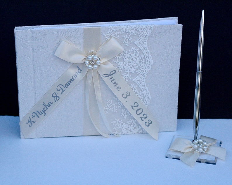 May include: A cream-coloured guest book with an embossed floral pattern, decorated with a cream satin ribbon, white lace trim, and a central pearl and rhinestone embellishment. The ribbon features the text K. Nycha & Damond and June 3, 2023.