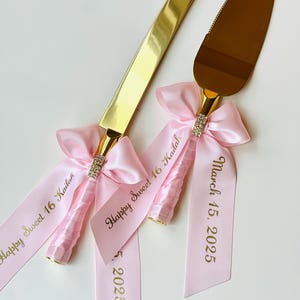 May include: Gold cake knife and server set with pink satin ribbons. The ribbons are tied with bows and have the text "Happy Sweet 16 Kailah" and "March 15, 2025" printed in gold.