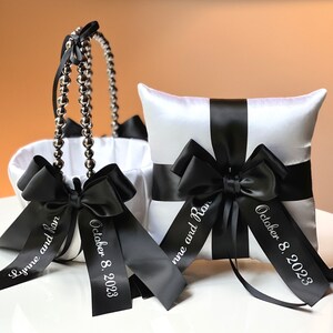 May include: White satin pillow with black satin ribbon and bow. The ribbon is personalized with the text "Lynne and Ron" and "October 8, 2023". A white satin flower girl basket with black satin ribbon and bow is also pictured. The ribbon is personalized with the text "October 8, 2023".