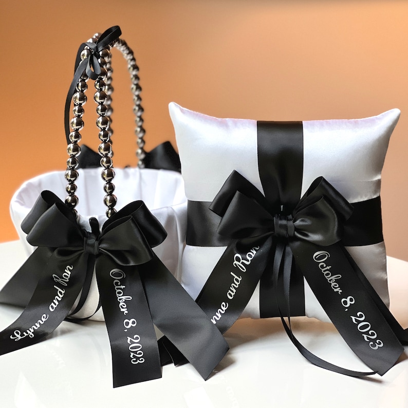 May include: White satin pillow with black satin ribbon and bow. The ribbon is personalized with the text "Lynne and Ron" and "October 8, 2023". A white satin flower girl basket with black satin ribbon and bow is also pictured. The ribbon is personalized with the text "October 8, 2023".