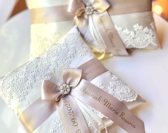 May include: Two ivory ring bearer pillows with lace trim and satin ribbons. Each pillow has a bow with a rhinestone embellishment and a ribbon printed with a name and date. Wedding decor.