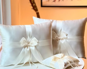 May include: Three white satin pillow covers with bows and a small white satin ring pillow with a bow and pearl accents. The pillows are arranged on a white table.