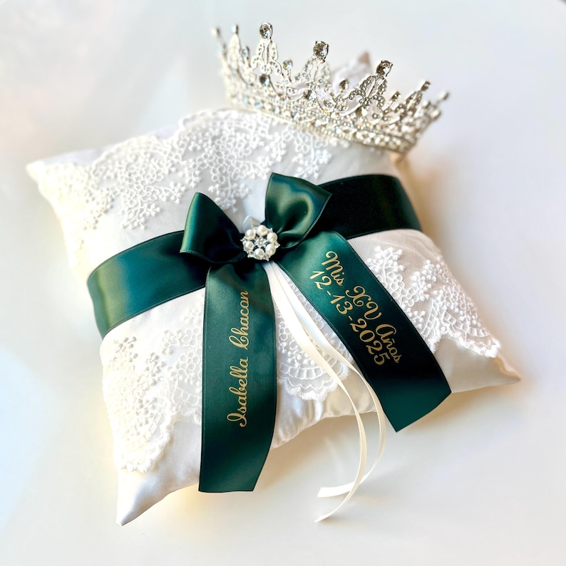 Peut inclure: Un coussin blanc en dentelle avec un ruban vert fonc&eacute; et une couronne argent&eacute;e. Le ruban est orn&eacute; d'un bijou et d'un texte dor&eacute; "Isabella Chacon" et "Mis XV A&ntilde;os 12-13-2025".