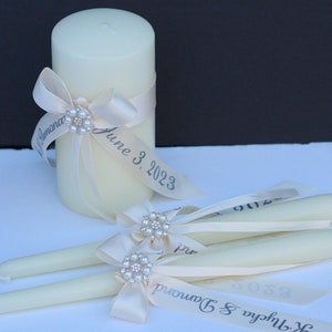 May include: A cream-coloured pillar candle and two matching taper candles, each adorned with a cream satin ribbon, a pearl and rhinestone embellishment, and printed text that reads "K. Nycha & Damond" and "June 3, 2023" in a dark grey font.