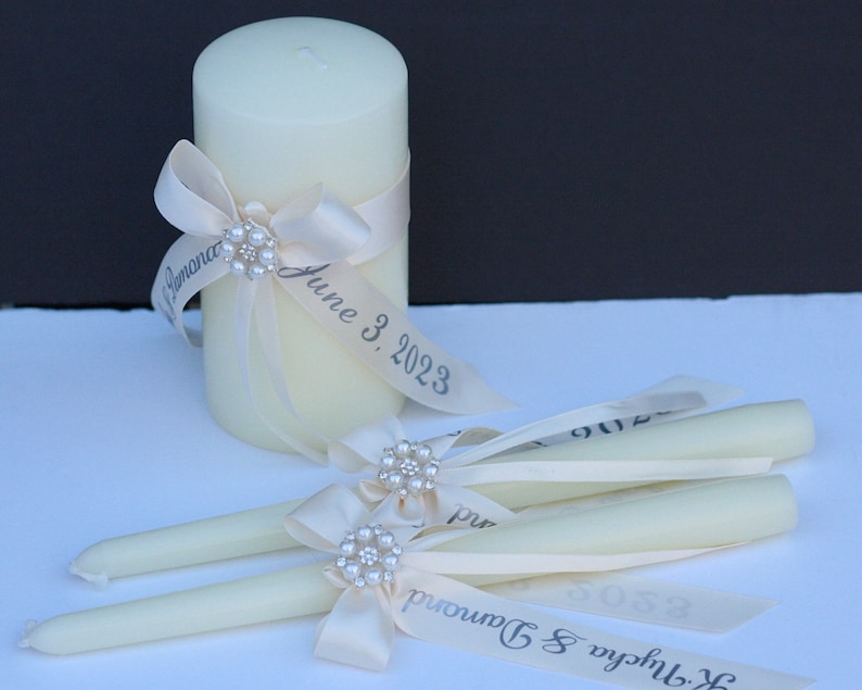 May include: A cream-coloured pillar candle and two matching taper candles, each adorned with a cream satin ribbon, a pearl and rhinestone embellishment, and printed text that reads "K. Nycha & Damond" and "June 3, 2023" in a dark grey font.