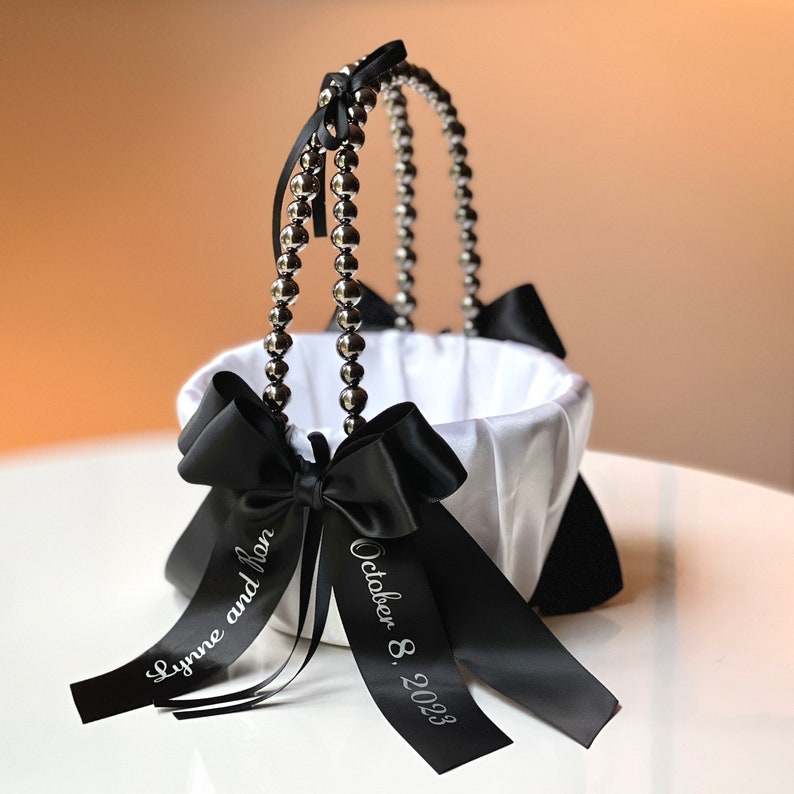 May include: White satin flower girl basket with a black satin bow and black ribbon with the text "Lynne and Ian October 8, 2023". The basket has a silver beaded handle.