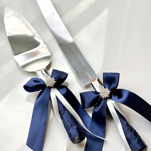 Puede incluir: Un juego de dos cuchillos para cortar pastel de plata con lazos de cinta de satén azul marino y blanco y detalles de diamantes de imitación.