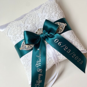 May include: White satin ring pillow with a teal satin ribbon tied in a bow. The ribbon is personalized with the text "Tiffany & Michael" and the date "06/23/2023".
