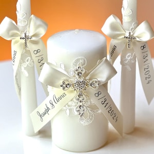 Puede incluir: Tres velas de unidad de boda blancas con cinta de satén marfil y una cruz enjoyada. La vela del centro tiene el texto "Joseph & Anna 8/31/2024" en la cinta.