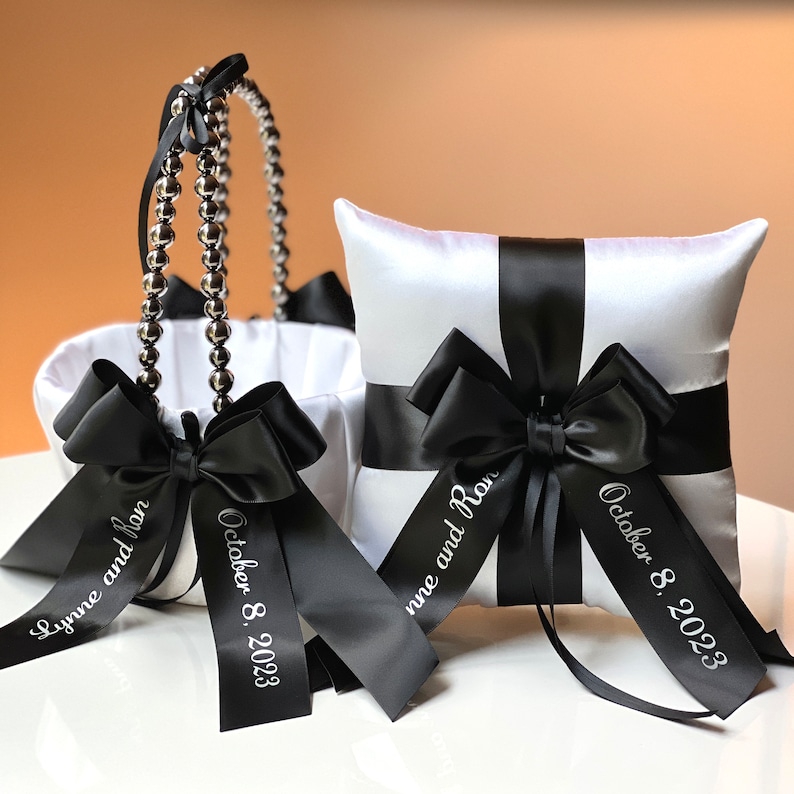 May include: White satin pillow with black satin ribbon and bow, and a white satin flower girl basket with black satin ribbon and bow. Both items have the text "Lynne and Ron October 8, 2023" printed on the ribbon.