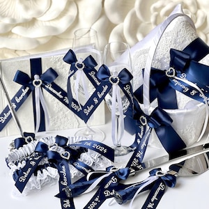Może przedstawiać: Zestaw akcesoriów ślubnych w kolorze białym i granatowym, w tym kieliszki do szampana, księga gości, serwer do ciasta i podwiązka. Każdy element jest ozdobiony granatowymi wstążkami, ozdobami w kształcie serca oraz spersonalizowanym tekstem "Delan & Carolee" i "December 7th 2024".