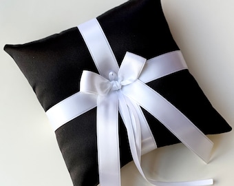 May include: A black pillow with a white satin ribbon tied in a bow. The pillow is designed for holding wedding rings.