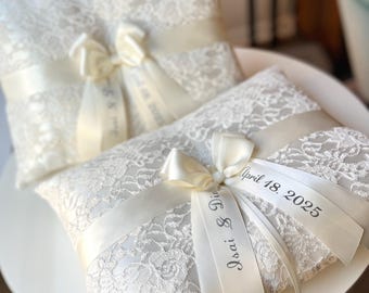 May include: Two ivory lace ring bearer pillows with cream-colored satin ribbons and bows. The pillows have the names "Isai & Dina" and the date "April 18, 2025" printed on the ribbons. The pillows are on a white surface.