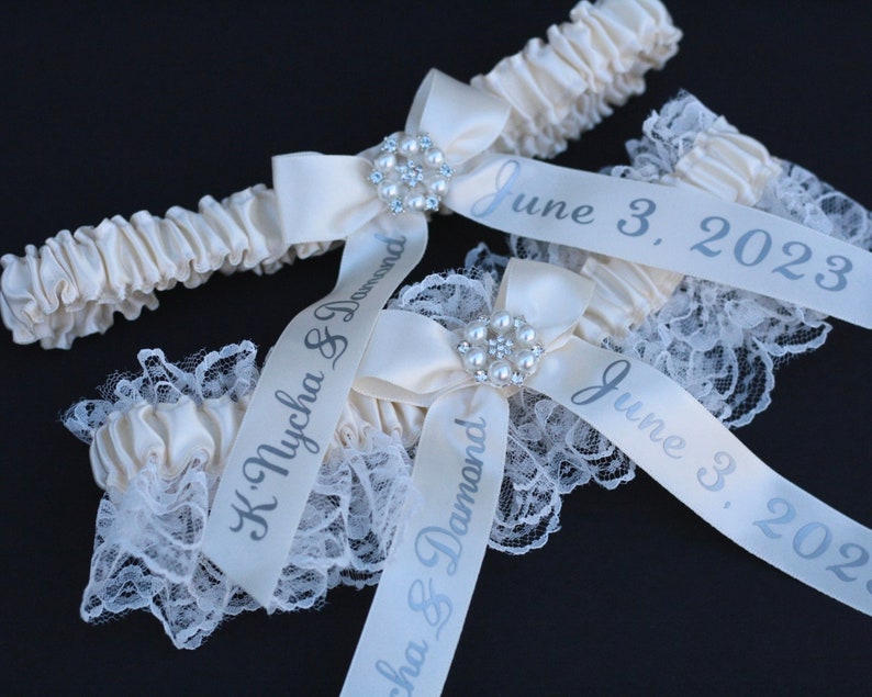 May include: Two ivory satin wedding garters featuring white lace trim, a central bow with a faux pearl and rhinestone embellishment, and printed silver text reading K. Nycha & Damond and June 3, 2023, on the hanging satin ribbons.