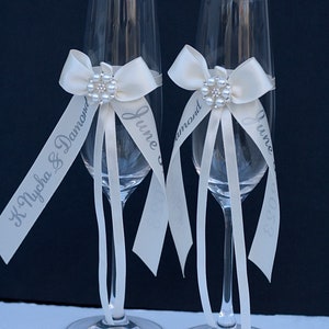 May include: Two clear glass champagne flutes, each adorned with a cream-coloured satin ribbon tied in a bow. A circular brooch featuring faux pearls and clear crystals sits at the centre of each bow. Long ribbon tails are printed with text.