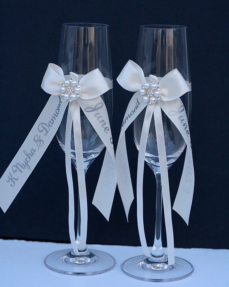 May include: Two clear glass champagne flutes, each adorned with a cream-coloured satin ribbon tied in a bow. A circular brooch featuring faux pearls and clear crystals sits at the centre of each bow. Long ribbon tails are printed with text.