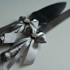 Può includere: Un set di coltello e server per torta argentati con fiocchi di nastro di raso grigio e dettagli in strass.